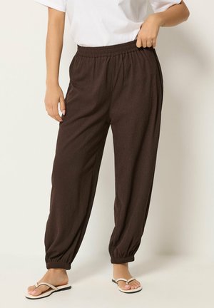Persoon draagt losse donkerbruine broek met textuur, elastische taille en boorden, gecombineerd met witte teenslippers en een witte top met korte mouwen.