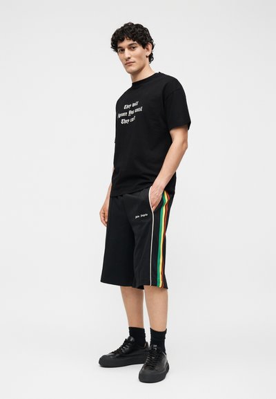 Άνδρας που στέκεται φορώντας μαύρο T-shirt με λευκό κείμενο και μαύρα σορτς με πράσινες, κίτρινες και κόκκινες πλευρικές ρίγες και μαύρα αθλητικά παπούτσια.