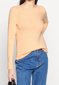 Ljus persikofärgad stickad tröja med båturringning och långa ärmar, kombinerad med blå denimjeans. Mjuk textur och avslappnad passform.