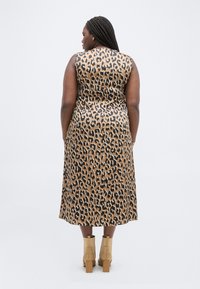 Γυναίκα με πλεξούδες, φορώντας αμάνικο, καφέ και μαύρο φόρεμα με λεοπάρ μοτίβο μήκους μέχρι τη μέση της γάμπας και ταμπά μποτάκια με τακούνι, στέκεται με την πλάτη γυρισμένη.