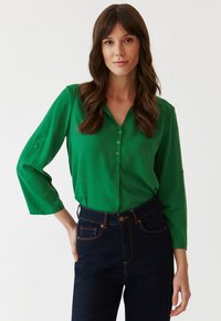 Bluză verde, ușoară, cu mâneci lungi și butoni, cu decolteu în V și tiv rotunjit, asortată cu blugi din denim bleu închis, cu talie înaltă.