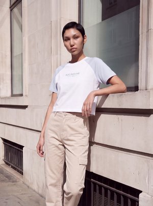Un mannequin pose devant un bâtiment en pierre, appuyé contre le rebord d’une fenêtre, avec un T-shirt blanc et un pantalon cargo beige.