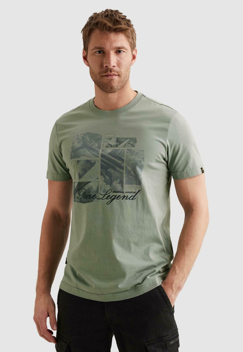 Άνδρας φοράει ελιάς πράσινο t-shirt με μαύρα γραφικά και κείμενο «PME Legend Original Aviator», στέκεται με το ένα χέρι στην τσέπη μαύρου παντελονιού cargo.