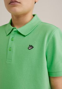 Žalias polo marškinėliai, pagaminti iš tekstūruoto audinio, su trimis mygtukais ir juodu siuvinėtu paukščio logotipu ant krūtinės.