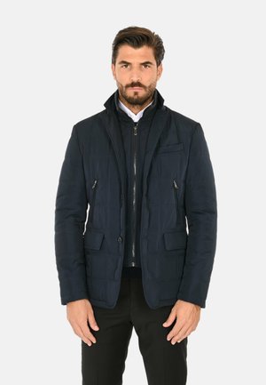 Uomo con capelli scuri e barba che indossa una giacca trapuntata nera sopra uno strato nero con cerniera e una camicia bianca con colletto su sfondo bianco.