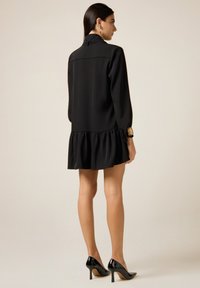 Zwarte jurk met een uitlopende zoom, lange mouwen en een hoge kraag. Gladde stof met een getailleerde pasvorm, gecombineerd met zwarte hoge hakken.