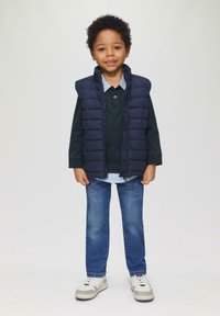 Gilet matelassé navy sur une chemise à manches longues vert foncé, col à carreaux bleu clair, jean bleu et baskets blanches avec des accents gris.