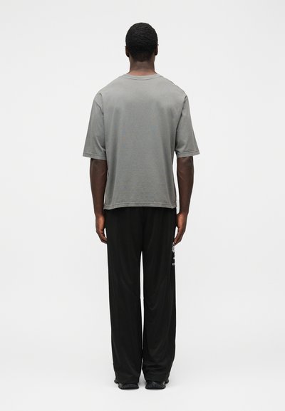 Šedé tričko s krátkym rukávom a voľným strihom, kombinované s čiernymi voľnými nohavicami, pohľad zozadu na jednoliatom pozadí.