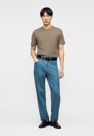 Άνδρας που φοράει καφέ κοντομάνικο πουκάμισο με υφή, μπλε τζιν με μαύρη ζώνη και μαύρα δερμάτινα παπούτσια, στέκεται με το ένα χέρι στην τσέπη.