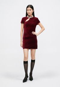 Burgundowa aksamitna sukienka z krótkimi rękawami, z bocznym wycięciem. W komplecie z czarnymi podkolanówkami i szpilkami z wąskim noskiem.