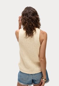 Beige gebreide mouwloze trui met een gestructureerde afwerking, gecombineerd met denim shorts en een gouden armband. Zacht, casual ontwerp.