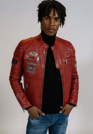 Veste de moto en cuir rouge avec des accents matelassés sur les épaules, plusieurs patchs brodés et des fermetures éclair, portée sur un col roulé noir.