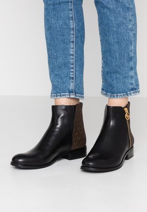 Personne portant des bottines en cuir noir avec des panneaux arrière marron à motifs et des détails de fermeture éclair dorée, associées à un jean bleu raccourci.