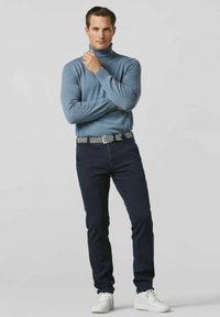 Helles blaues Rollkragensweater, marineblaue Jeans, gemusterter Gürtel und weiße Sneakers. Modell steht mit verschränkten Armen vor einem neutralen Hintergrund.