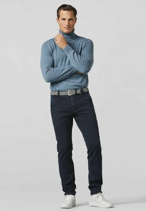 Lichtblauwe coltrui, marineblauwe jeans, stoffen riem en witte sneakers. Model staat met gekruiste armen, neutrale achtergrond.