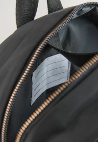 Czarna torba na plecy wewnątrz z gładką podszewką i zamykaną na zamek kieszonką z etykietą na dane kontaktowe. Widoczne teksturowane czarne paski.