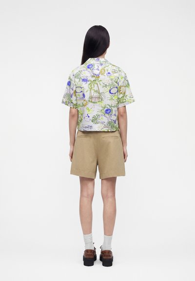 Γυναίκα με μακριά σκουρόχρωμα μαλλιά φορά κοντομάνικο, πολύχρωμο πουκάμισο με σχέδια και μπεζ σορτς μέχρι τα γόνατα, στέκεται γυρισμένη πλάτη σε λευκό φόντο.