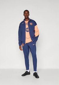 Muž v námorníckej a broskyňovej súprave FC Barcelona Nike s čiernymi topánkami, stojaci na pozadí svetlošelej farby.