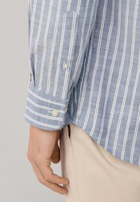Blauw-wit gestreepte stoffen shirt met knoopmanchetten en een ronde zoom, gemaakt van lichtgewicht materiaal, met een close-up van de mouw en hand.