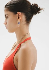 Aros de oro en forma de aro con acento azul, collar de cadenas en capas, blusa sin mangas roja, piel suave y cabello recogido, mostrados de perfil.