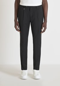 Zwarte op maat gemaakte broek van gladde stof met platte voorplooien en een discrete sluiting. Gepaard met witte sneakers.