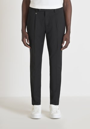 Pantalones negros ajustados de tejido suave con pliegues planos al frente y un cierre discreto. Combinados con zapatillas blancas.