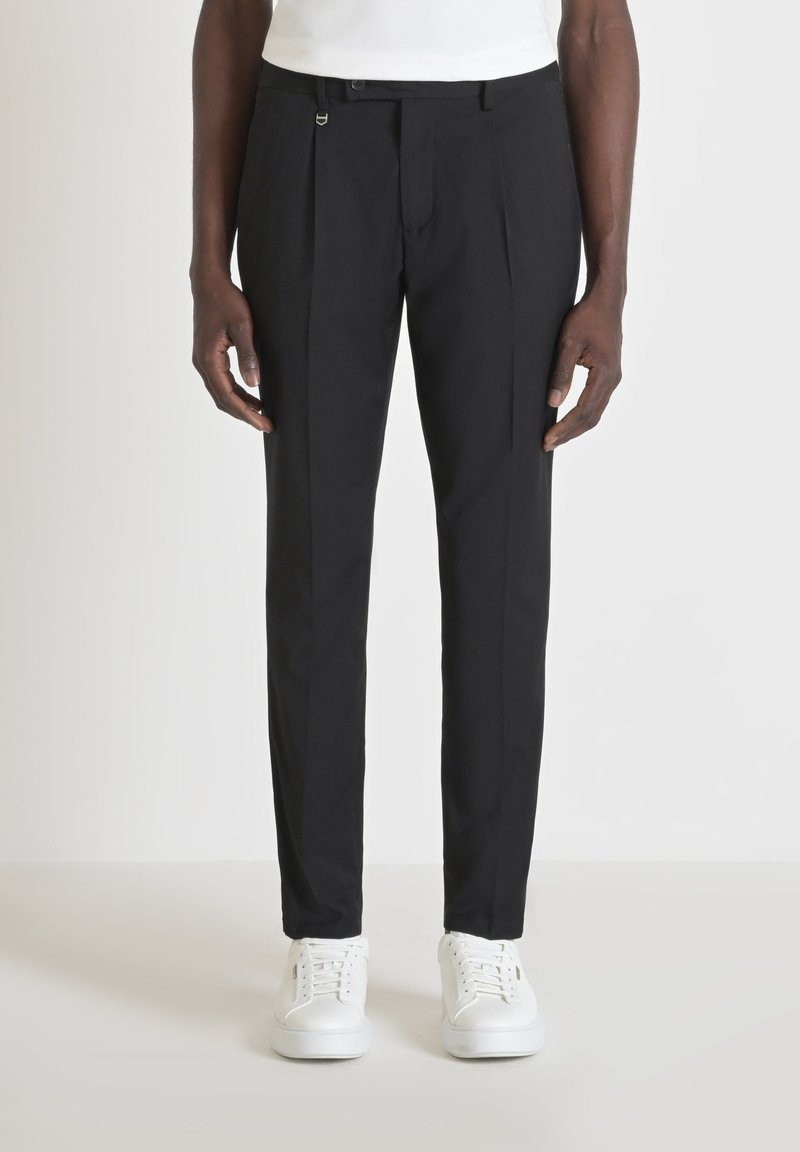 Zwarte op maat gemaakte broek van gladde stof met platte voorplooien en een discrete sluiting. Gepaard met witte sneakers.