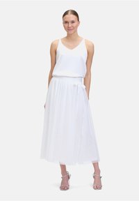 Weißes ärmelloses Top mit V-Ausschnitt, kombiniert mit einem langen, fließenden weißen Rock mit gebundenem Taillenbund und silbernen Riemchen-High-Heels.
