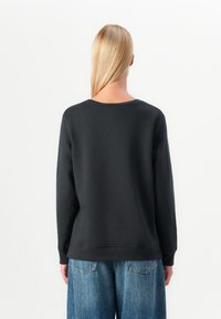 Pitkähiuksinen henkilö, jolla on pitkä vaalea tukka ja joka on kuvattu selin, pukeutuneena mustaan pitkähihaiseen paitaan ja sinisiin farkkuihin, yksinkertaista valkoista taustaa vasten.