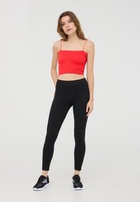 Czerwony prążkowany top na krótki rękaw z cienkimi ramiączkami, w połączeniu z czarnymi elastycznymi legginsami oraz czarnymi butami sportowymi z białymi podeszwami. Jasne tło.