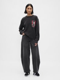 Μαύρο φαρδύ φούτερ με γραφικό σχέδιο στο στήθος, συνδυασμένο με μαύρο τζιν παντελόνι φαρδιάς γραμμής με διαγώνια ραφή. Μαύρα αθλητικά παπούτσια.