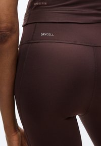 Κοντινό πλάνο ατόμου που φοράει αθλητικό κολάν σκούρου καφέ με ορατά λογότυπα «DRYCELL» και «CLOUDSPUN» στη μέση και το ισχίο.