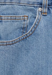 Gros plan sur la poche d'un jean en denim bleu clair avec des coutures orange, une boucle de ceinture et un rivet en métal.