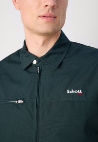 Donkergroene lichtgewicht jas met een staande kraag, voorzak met rits en geborduurd 'Schott NYC' logo in wit en rood. Soepele textuur.