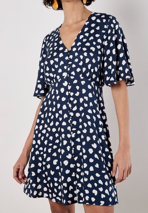 Vrouw draagt een marineblauwe jurk met witte, onregelmatige hartvormige vlekken, knopen aan de voorkant en korte, wapperende mouwtjes.