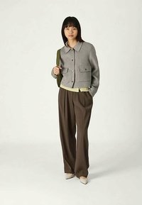 Veste grise courte avec deux poches avant, superposée sur un haut vert clair, associée à un pantalon large marron et des chaussures beiges à bouts pointus.