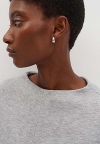 Orecchino a goccia argentato su un taglio di capelli corto. La persona indossa un maglione grigio chiaro, texture, con scollatura rotonda.