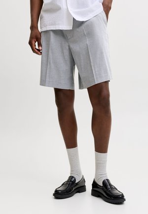 Hombre que lleva pantalones cortos plisados de color gris claro, calcetines blancos, mocasines negros y una camisa blanca con una mano en el bolsillo frente a un fondo liso.