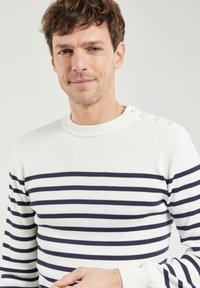 Maglione bianco a righe con strisce orizzontali blu navy, texture a coste e bottoni sulle spalle. Design aderente con scollo rotondo.