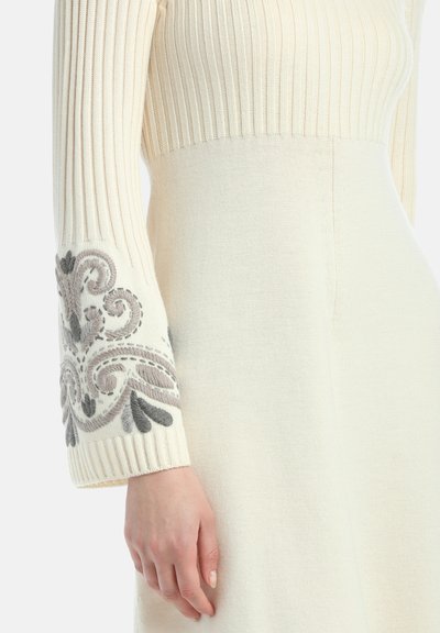 Pogled v bližino belega puloverja z rebrastim rokavom, okrašenega z sivo vezenimi cvetličnimi vzorci, ob sproščeni roki, ki leži poleg pletenih belih kril.