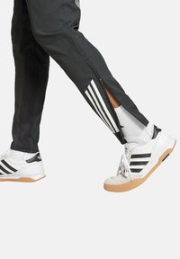 Schwarze Sporthosen mit Reißverschlüssen an den Knöcheln und weißen Streifen. Getragen mit weißen und schwarzen Sneakers mit einer Gummisohle und strukturierten Akzenten.