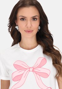Vit kortärmad t-shirt med en stor rosa rosett-grafik på framsidan. Modellen bär överdimensionerade örhängen i silver och pärlor.