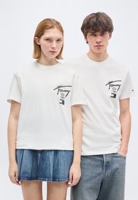 Δύο νέοι ενήλικες που φορούν λευκά τυπωμένα T-shirts Tommy, η γυναίκα με φούστα από τζιν και ο άνδρας με τζιν παντελόνι, στέκονται μπροστά σε ένα απλό φόντο.