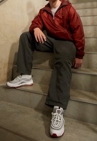 Röd lättviktsjacka, grå byxor och vita Nike Air Max 97 sneakers med röda accenter, sittande på betongtrappor.