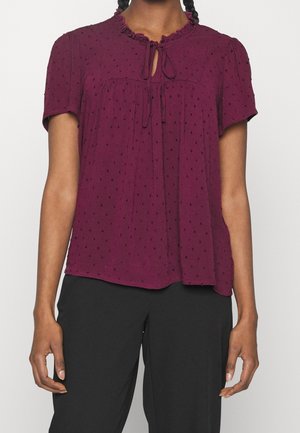 Vrouw draagt een bordeauxroze blouse met korte mouwen en een bindstrik bij de hals, gecombineerd met losse zwarte broek, staand tegen een witte achtergrond.