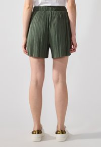 Shorts plissés verts avec une taille élastique, longueur mi-cuisse et texture lisse. Portés avec des chaussures blanches avec des accents dorés.