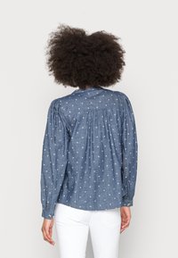 Džínová košile s kulatým výstřihem, dlouhými nabíranými rukávy, nařaseným zadním dílem a bílými křížovými vzory na modrém pozadí.