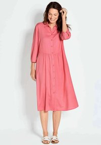 Vestito rosa con pulsante che arriva fino al ginocchio, maniche a tre quarti, vita arricciata e orlo che si ferma al ginocchio. Tessuto liscio con una leggera texture.
