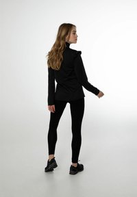 Zwarte blouse met lange mouwen en ruches op de schouder, gecombineerd met zwarte leggings en sportschoenen, gezien van achteren.