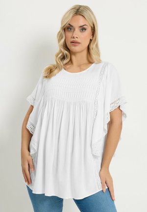 Kvinde med langt blondt hår iført en løs hvid bluse med blonderdetaljer og blå jeans, stående foran en ensfarvet lys baggrund.